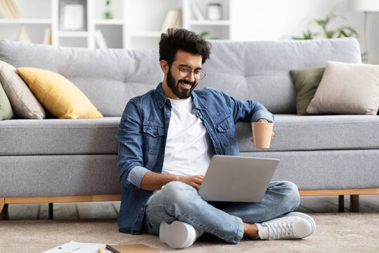 Smiling Indian man using laptop while sitting cross-legged on floor at home