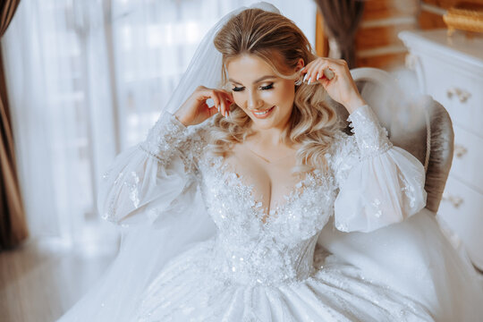 fashion photo of a beautiful bride with blond hair in an elegant wedding dress and stunning makeup in the room on the morning of the wedding. The bride is preparing for the wedding