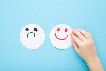 Baby hand with bad and happy smileys on light blue table background. Pastel color. Negative and positive smiling face expressions on white paper. Closeup. Children mood concept. Top down view. - obrazy, fototapety, plakaty
