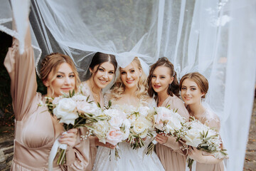 Wedding photography in nature. A blond bride and her girlfriends pose under a veil, holding bouquets