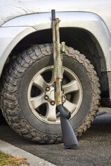 A camouflage-colored shotgun gun standing against a mud-covered wheel of a silver SUV