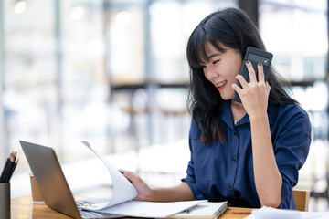 Smiling Asian businesswoman talking on the phone while working with laptop in office