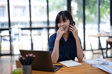 Obraz premium Smiling Asian businesswoman talking on the phone while working with laptop in office