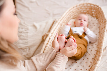 Fototapeta na wymiar Woman holding baby boy feet laying in baby bassinet in bed close up. Motherhood.