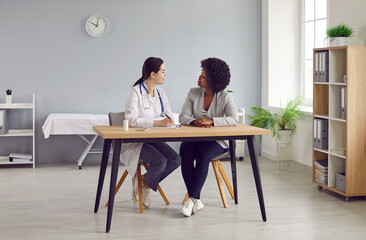 Female doctor and patient communicating in medical room at clinic. Young physician sitting at desk together with African American woman, writing out prescription and explaining how to take medicine - obrazy, fototapety, plakaty