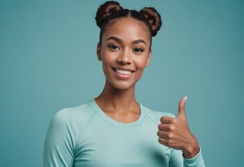A woman with hair in buns, making a friendly thumbs up gesture in active wear. Studio shot with a blue background. - obrazy, fototapety, plakaty