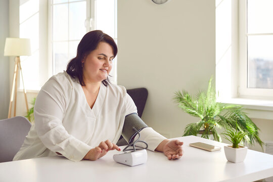 Young overweight woman with hypertension doing a self checkup at home. Fat lady sitting at a desk, using a modern digital sphygmomanometer, and taking her blood pressure and heartbeat rhythm herself