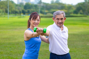 Senior asian man is using dumbbell for weight training strength while his daughter is supporting in...