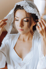 Fototapeta premium portrait of an incredibly beautiful girl bride in a white robe in the bedroom, the bride poses holding her veil in her hands and covers herself with it.
