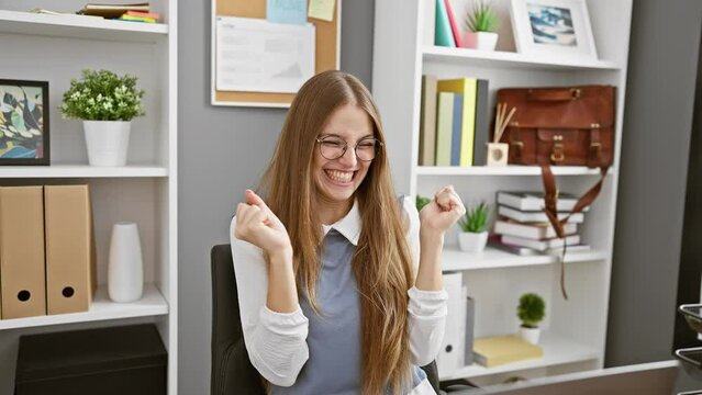 Ecstatic young blonde business woman at office, arms aloft in winning victory. her jubilant 'yes!' echoes triumph, celebrating her success with infectious joy
