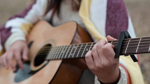 Close up of girl playing the guitar with capo outdoors on autumn nature. Girl covered in warm plaid play acoustic guitar outdoors