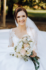 The brunette bride, smiling sincerely, sits on the ground in her unfolded dress and holds a...