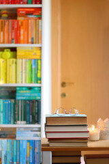 Stack of books, reading glasses and lit candles on the table. Rainbow bookshelf in the background. Selective focus.