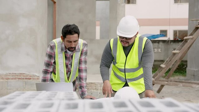 Male supervisor explains construction plan to junior engineer. Asian engineers in yellow vests work in team with laptop and document on table outdoor in front unfinished cement building, bricks
