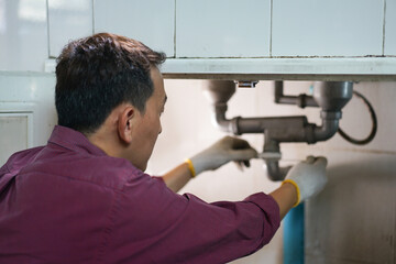 A male plumber repairs a pipeline or drain under the sink in kitchen room.