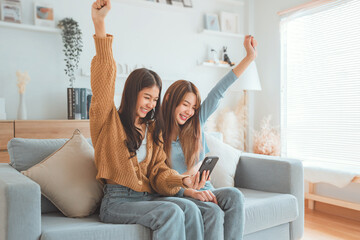 Exciting young asian women LGBT happy couple sitting on sofa using mobile phone in living room at...