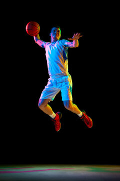 Athlete man basketball player passing ball in mid-air in action against black studio background in mixed neon light. Concept of professional sport, energy, strength and power, match, championship.