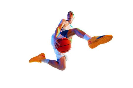 Low angle view. Athletic man in mid-jump shot with basketball ball against white studio background in neon light. Concept of professional sport, energy, strength and power, championship, tournament.