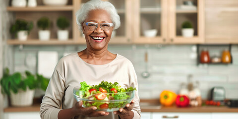 African-American old retired lady woman smiling happily and holding a healthy vegetable salad bowl on blurred kitchen background. Senior healthy lifestyle living eating habits long life concept - obrazy, fototapety, plakaty