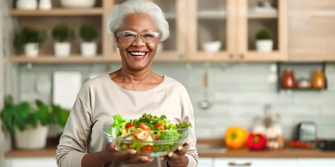 Poster African-American old retired lady woman smiling happily and holding a healthy vegetable salad bowl on blurred kitchen background. Senior healthy lifestyle living eating habits long life concept © Valeriia