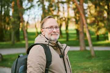 Outdoor portrait of handsome middle age man in green park, wearing warm jacket and backpack - 741618810