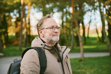 Outdoor portrait of handsome middle age man in green park, wearing warm jacket and backpack - 741618813