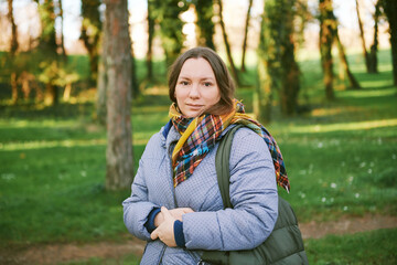 Outdoor portrait of 40 - 45 year old woman, wearing warm jacket and scarf, posing in green park - 741618833