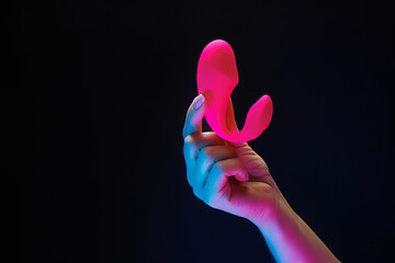 Close-up of a girl holding a red-colored sexual entertainment toy. Sex toy clitoral vibrator on a black background with neon lights. - obrazy, fototapety, plakaty