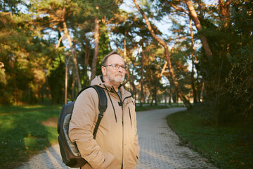 Outdoor portrait of handsome middle age man in green park, wearing warm jacket and backpack - 741618873