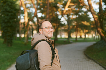 Outdoor portrait of handsome middle age man in green park, wearing warm jacket and backpack - 741618877