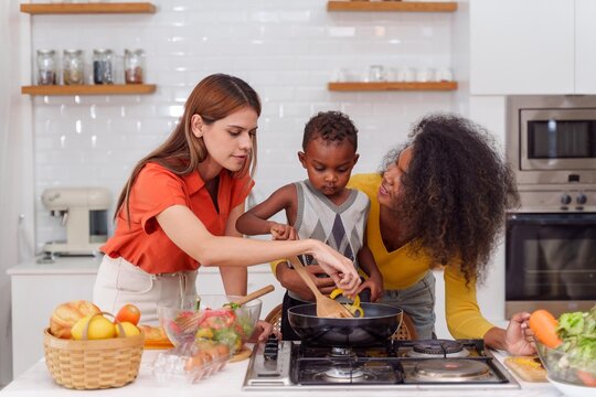 Happy Two asian women lgbtq lesbian and son making salad while preparing food in the kitchen having fun, mother and son cooking activity concept.