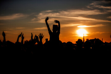 An unrecognizable group of people waves their hands at a concert or public event against the backdrop of the setting sun. Night music festival.