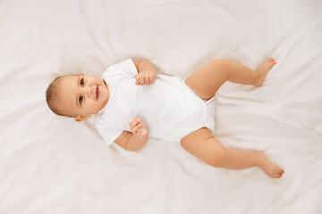Happy, smiling child, baby, adorable girl in white onesie lying on bed and lookin upwards against white background. Concept of childhood, family, care, motherhood, infancy, heath