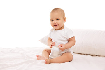 Little baby child, girl, adorable infant in white onesie sitting on bed with pillow and smiling against white background. Concept of childhood, family, care, motherhood, infancy, heath