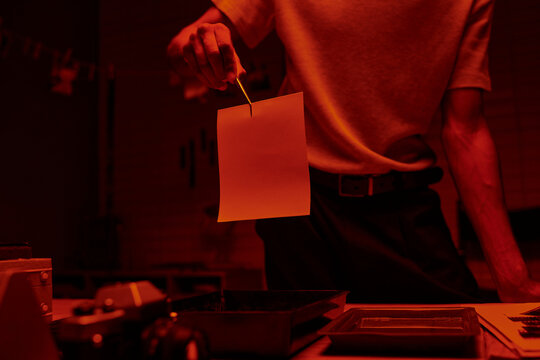 cropped shot of photographer holding tweezers with photo paper in a darkroom with red light