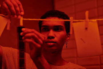 african american photographer hangs freshly developed film strip  in a red-lit darkroom, timeless