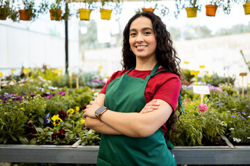A young, Latina woman works inside a nursery dressed in a green work apron. The dark-skinned woman...