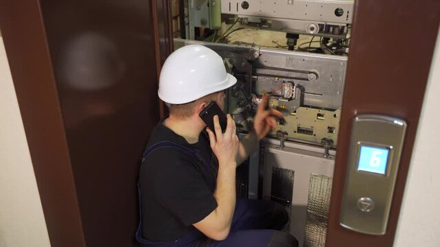 Emergency situation in elevator equipment. An engineer, worker, electrician near an open elevator shaft talks on the phone with technical support. Verifies performance indicators before repairs.