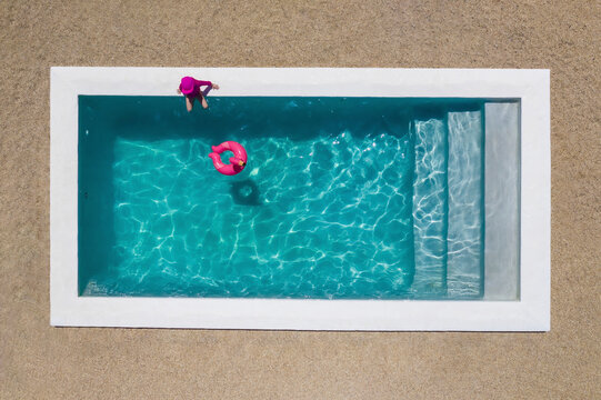 Aerial view of a girl wearing a pink costume sitting by a pool with clear blue water and a pink flamingo floatie in the pool, Cape Town, Western Cape, South Africa.