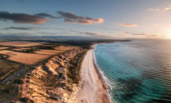 Aerial view of a white sandy beach edged with limestone cliffs and blue water at sunset with some clouds in the sky and farm fields at the top of the cliffs, Port Willunga, South Australia, Australia.
