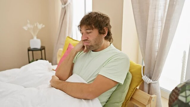 Handsome young caucasian man, bored and tired, wearing pyjamas, looking upset and thinking about depression problems indoors. he has crossed arms and a lazy expression in a bedroom setting.