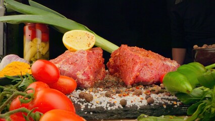 Chef cuts fresh meat on a wooden board on a dark background with vegetables and spices for marinade. Recipes and fresh produce concept.