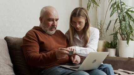 A pretty young woman shows her older mature father how to use a bank card and computer at home. An adult daughter teaches laptop apps to a focused middle-aged dad using modern technology.