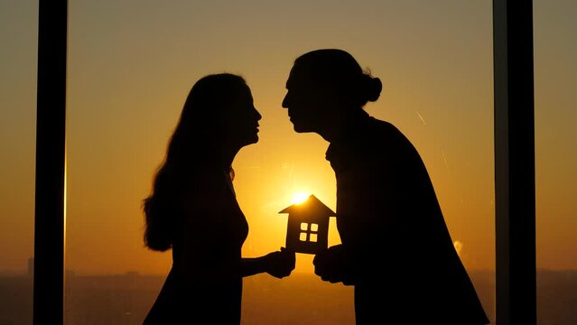 Silhouettes of young couple, facing each other by window, hold tiny paper house. Intimate moment, man leans forward, bringing his face close to woman, and they share gentle kiss.