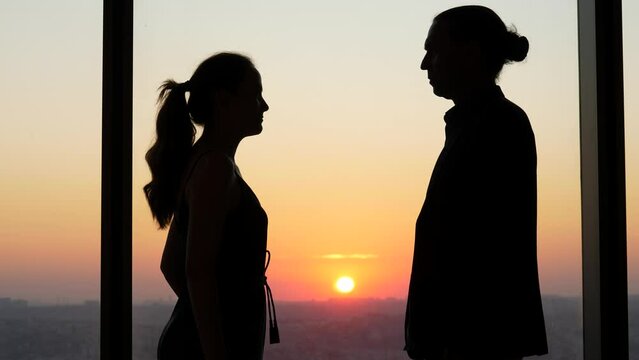 Couple stands in silhouette against window, facing each other. Tension is palpable, expressions strained. Their disagreement creating difficult moment, atmosphere is intense, standoff between two