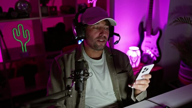 A young bearded man in a cap holding a smartphone podcasting with a microphone in a neon-lit gaming room at night.