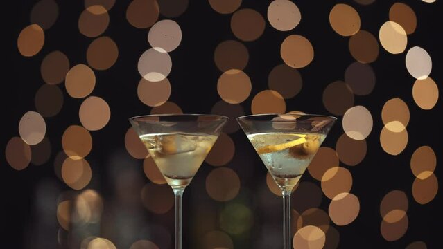 dos copas con martini, aparecen dos manos para tomarlas y brindar. La mano pertenece a una mujer y a un hombre. Fondo de luces bokeh desenfocadas.