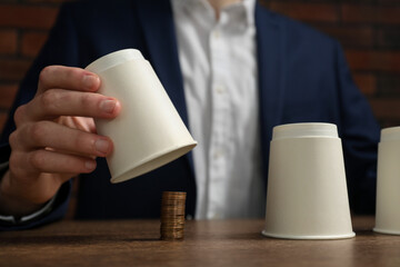 Shell game. Man showing stack of coins under cup at wooden table, closeup