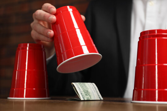 Shell game. Man showing dollar banknote under cup at wooden table, closeup