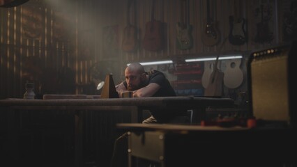 Tired carpenter looks at metronome, sits in workshop for making musical instruments. Male artisan,...